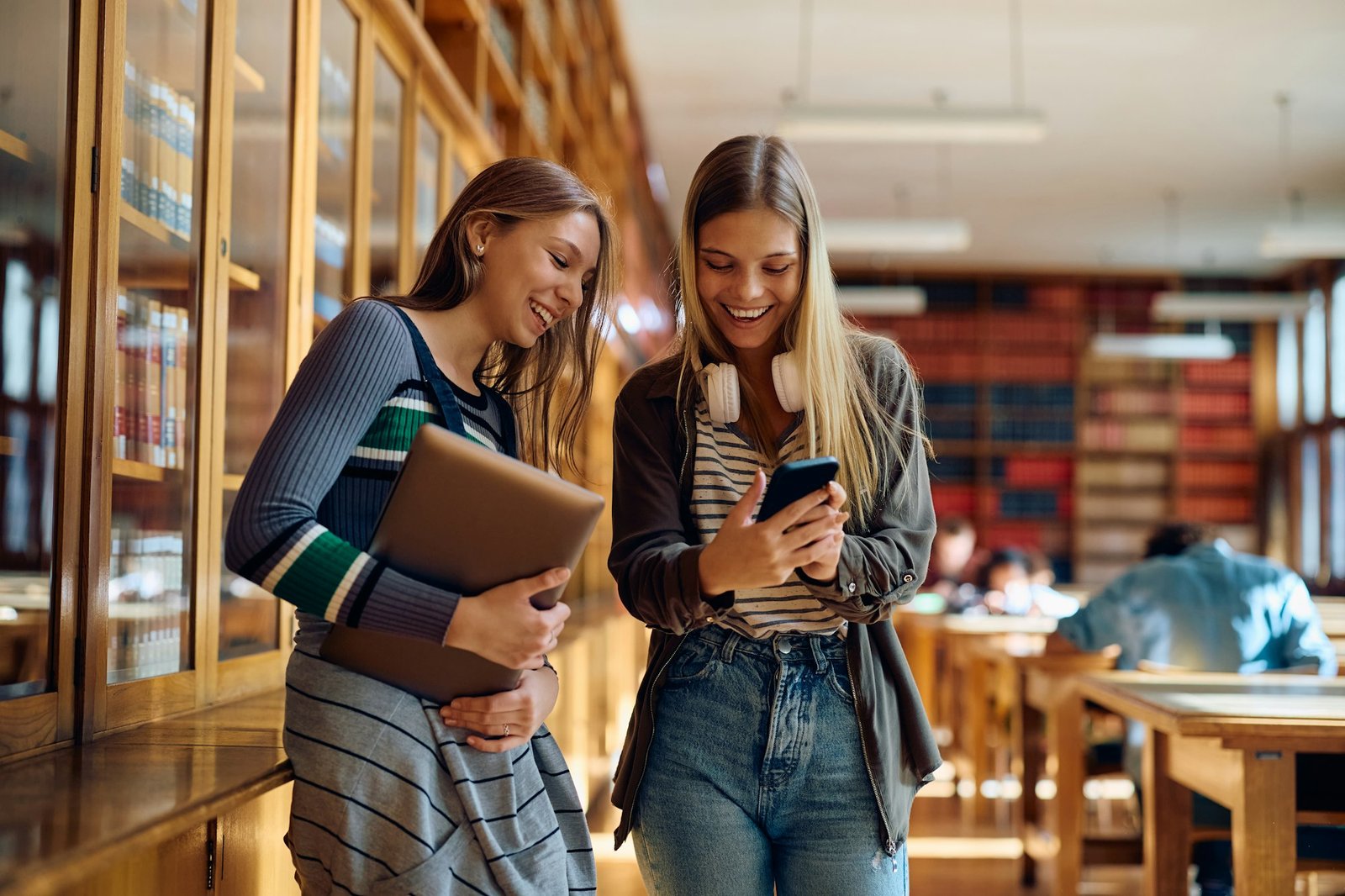 Happy teenage girls using mobile phone at high school library.